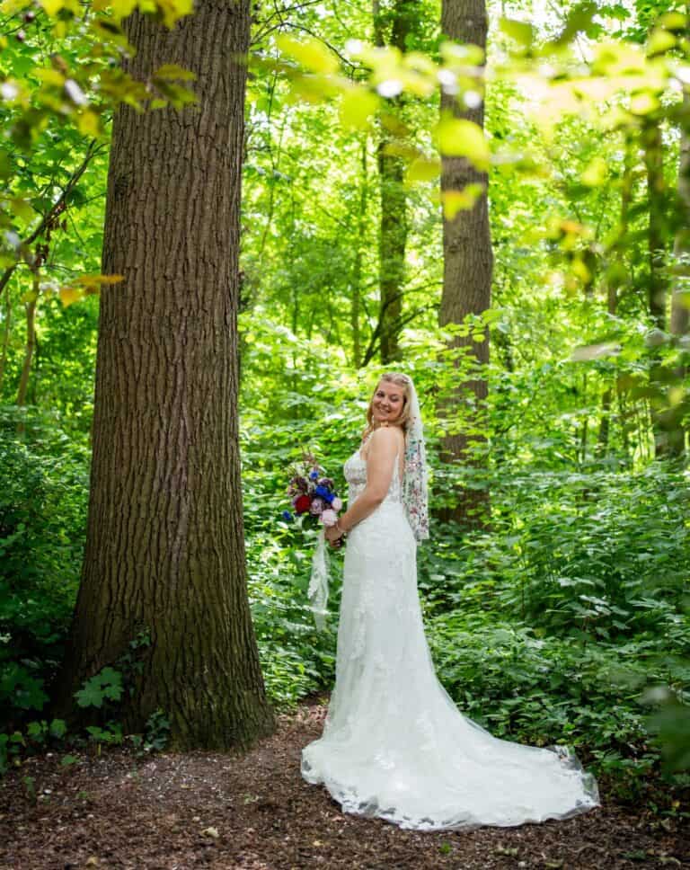 Een bruid in een bosrijke omgeving met groene bladeren en bomen, in een witte trouwjurk.