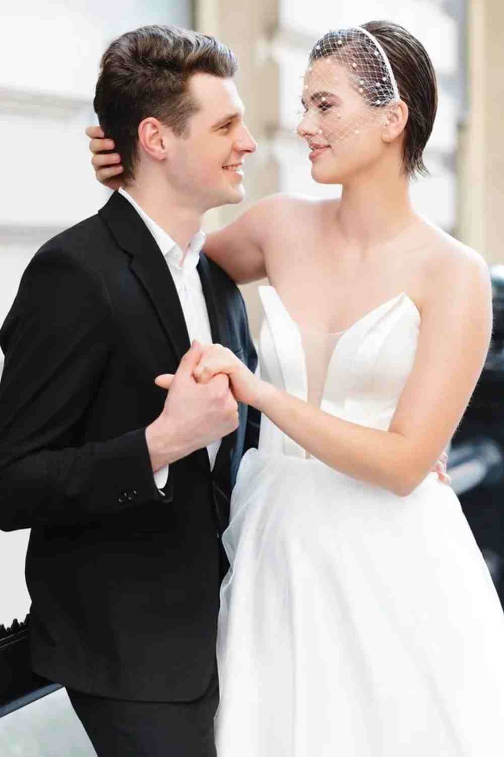 Bruid met slicked back los kort haar. Bruid draagt een birdcage sluier en staat met haar bruidegom