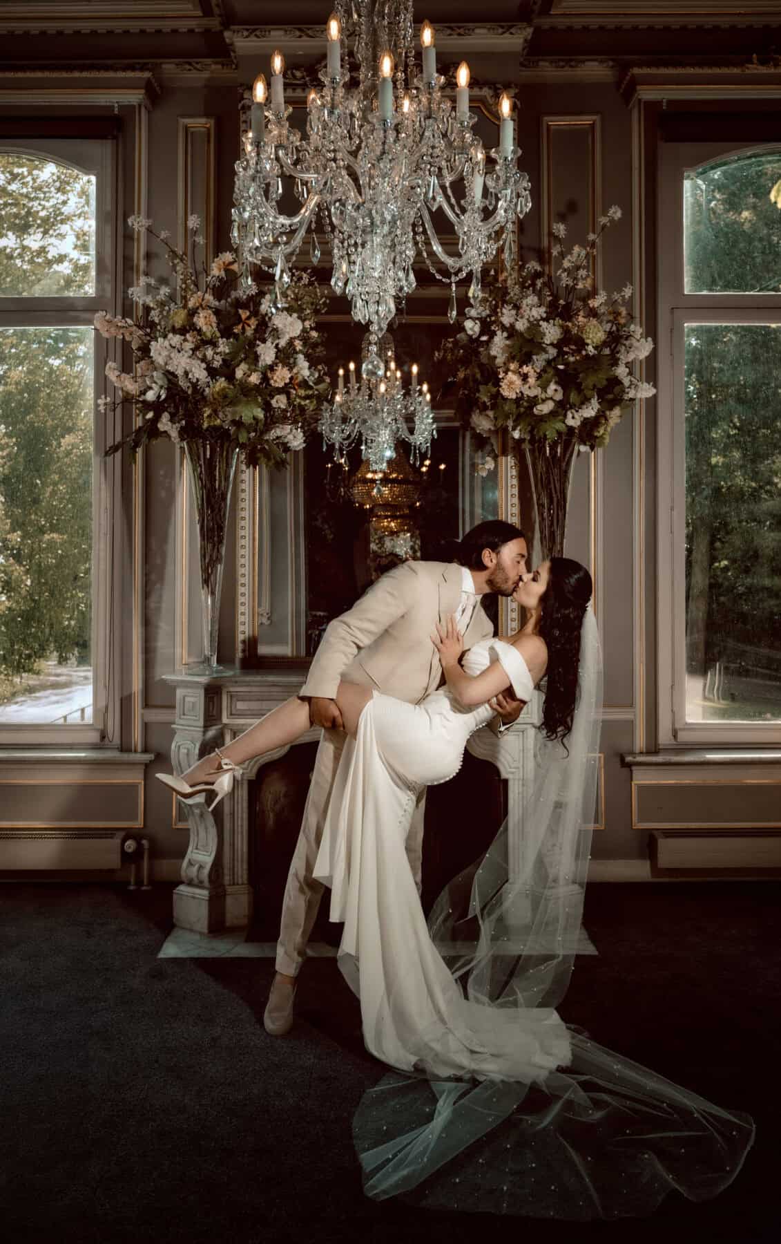 Huwelijkspaar danst onder chandelier in elegante trouwzaal met bloemen decor.