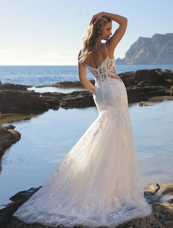 Honeymoonshop trouwjurk Goddess Azuree op strand met zee en rotsen.