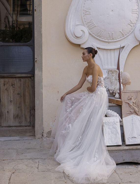 Elegante bruid in een romantische trouwjurk met kant en tule, zittend buiten voor een vintage muur.